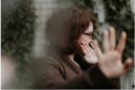 A person with glasses facing sideways, covering their face with one hand. The background features green foliage.