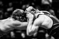 Two wrestlers in a match, wearing singlets and headgear, engaged in a grappling position. The image is in black and white.