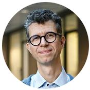 Stefano 175 Headshot of a person with curly hair, wearing glasses and a light-colored shirt, smiling slightly, with an indoor blurred background.