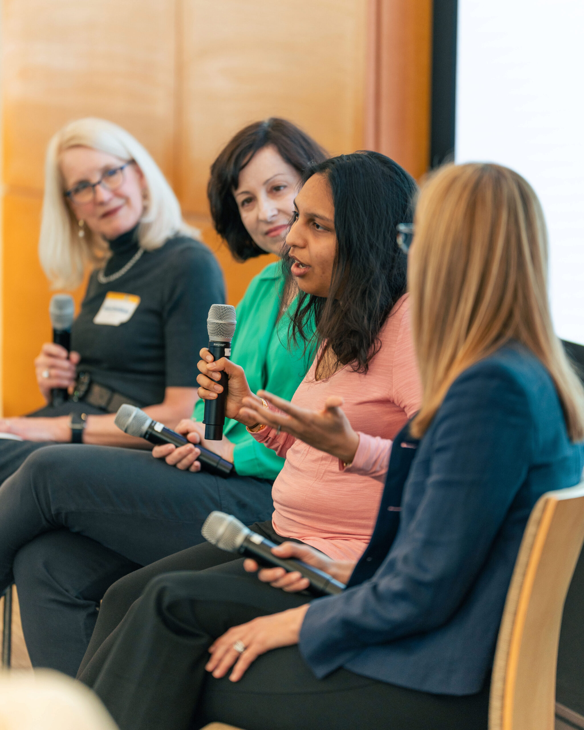 Four people in a panel discussion, each holding a microphone. They are seated in a line, engaged in conversation, in what appears to be a professional setting.