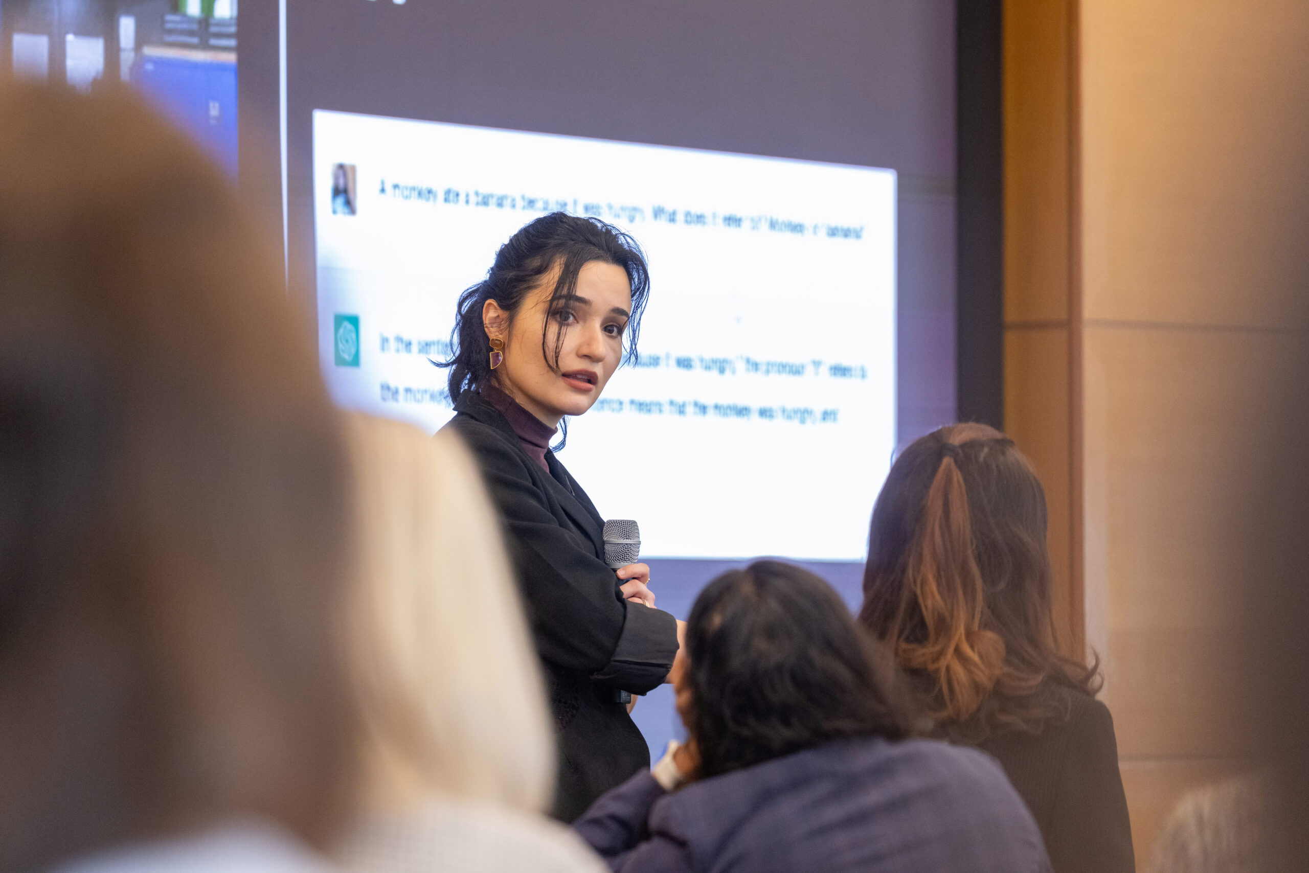 A person holding a microphone is giving a presentation in front of an audience. A projection screen displays text related to a sentence about a monkey eating a banana.