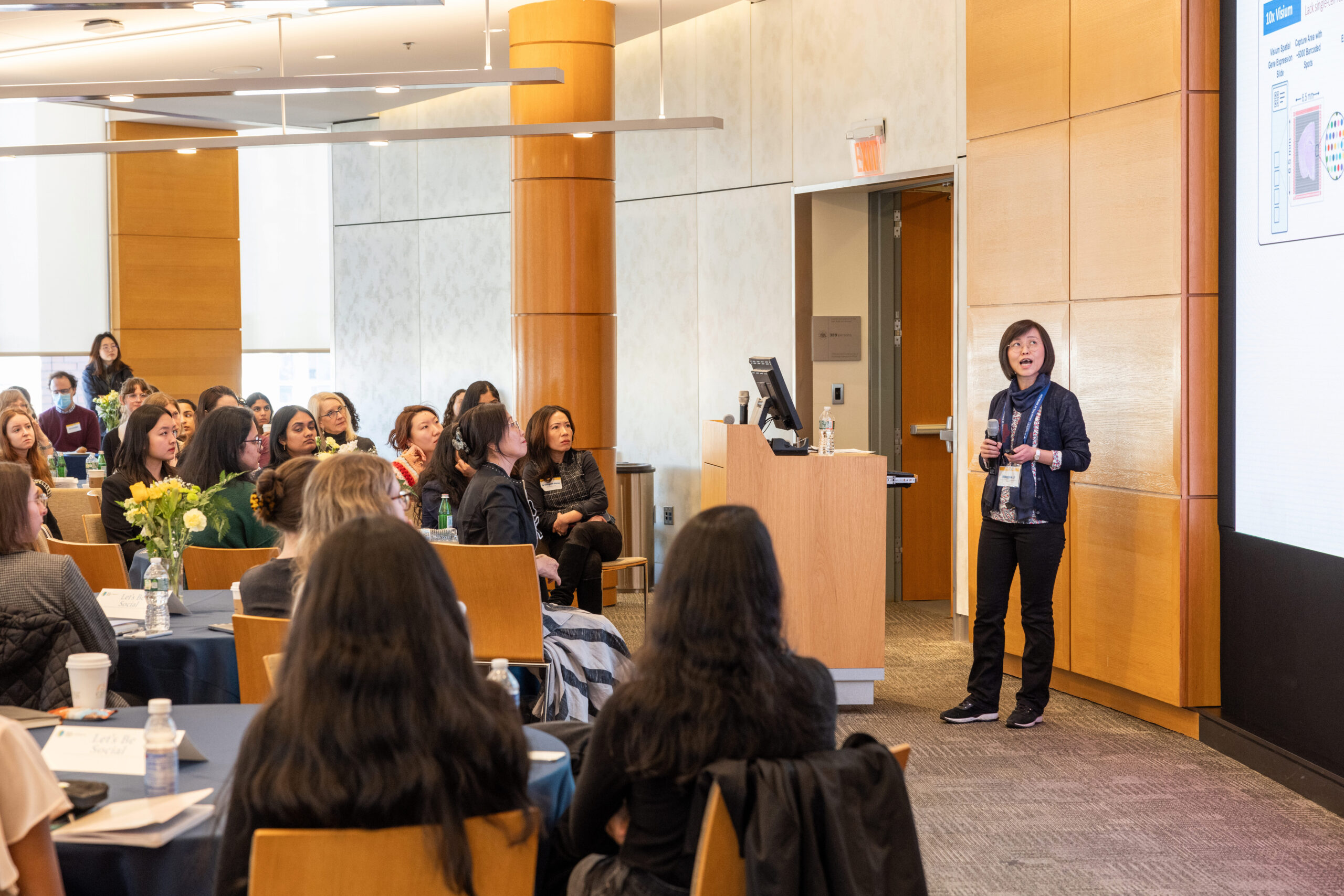 A presenter stands by a screen giving a presentation to a seated audience in a conference room. Attendees appear attentive, with tables in front of them.