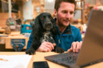 Carpenter With Pet Spaniel Working In Woodwork Workshop Using Laptop Together