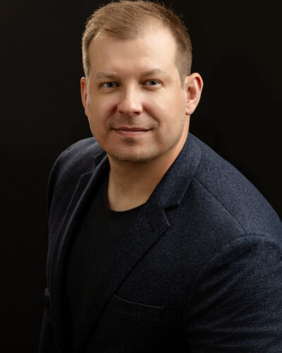 A person wearing a dark blazer and shirt is posing for a portrait against a dark background.