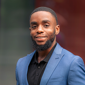 Headshot of a person with a beard wearing a blue blazer and black shirt, smiling against a blurred background.