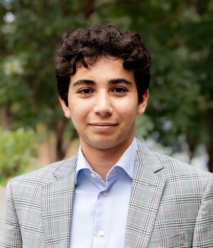 Headshot of a person with curly hair, wearing a plaid blazer and light blue shirt, standing outdoors with trees in the background.