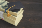 A stack of U.S. hundred-dollar bills topped with a graduation cap, symbolizing the cost of education or student debt.