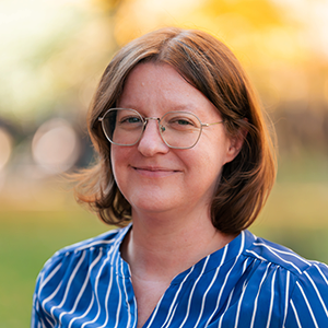 A headshot of a person with brown hair and glasses, wearing a blue and white striped shirt. The background is softly blurred, suggesting an outdoor setting.