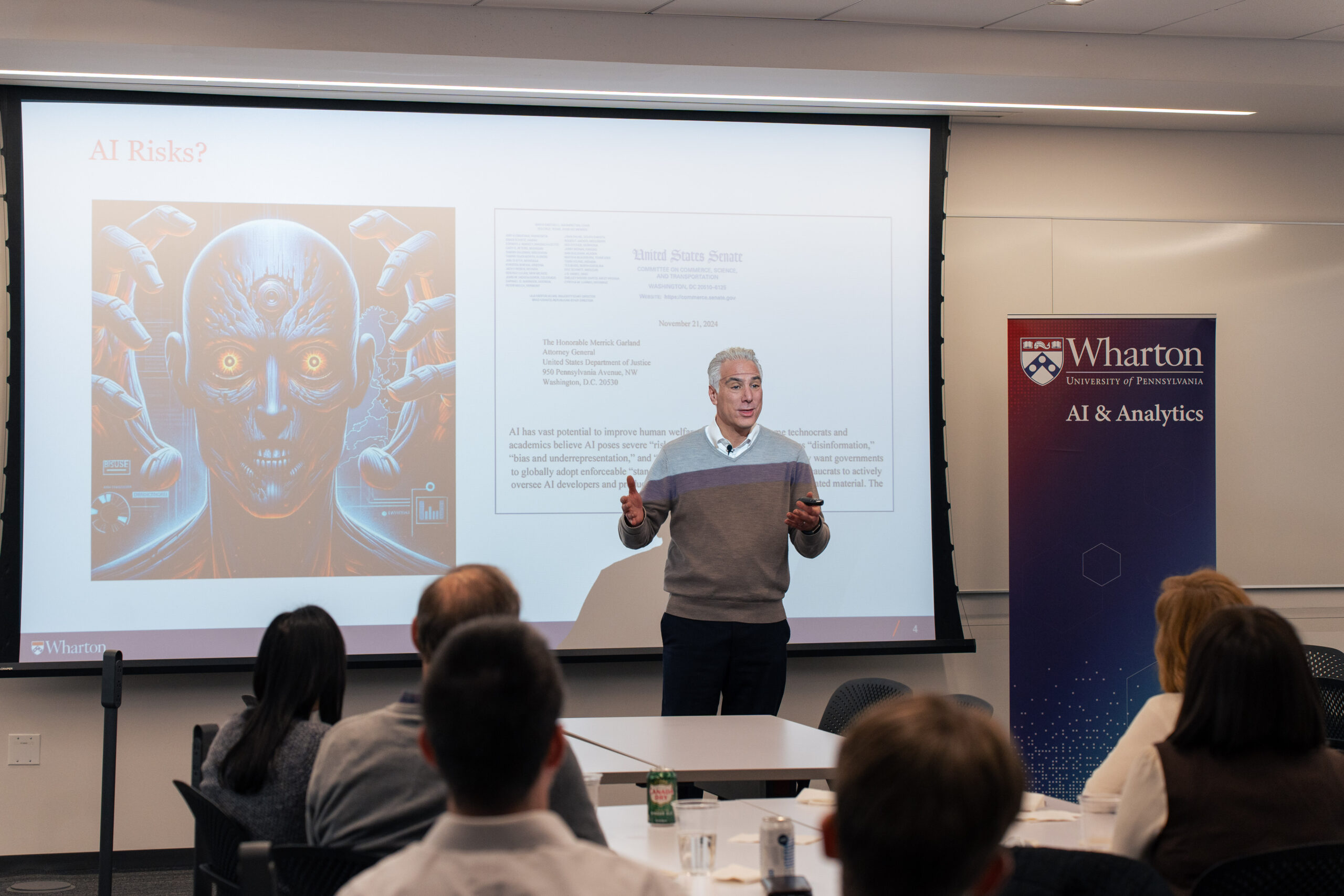 A presenter stands in front of an audience, giving a talk on AI risks. A projection screen displays an image of a menacing cyborg face and text related to AI concerns. A sign for "Wharton AI & Analytics" is visible.