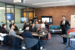 A person in formal attire is giving a presentation to a small group seated in a modern conference room. There is a brick wall and a screen displaying content in the background.