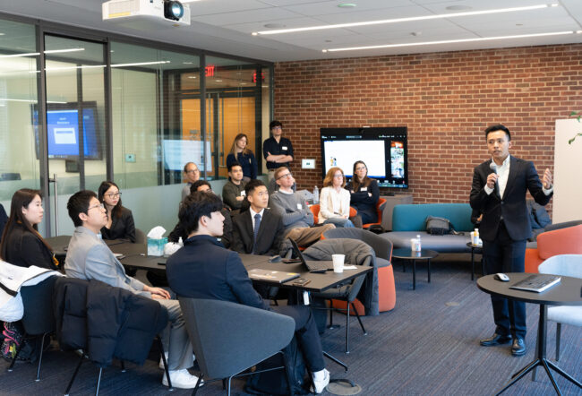 A person in formal attire is giving a presentation to a small group seated in a modern conference room. There is a brick wall and a screen displaying content in the background.