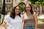 Two individuals smiling outdoors, standing in front of an arched building entrance. They are casually dressed in a white t-shirt and a beige top with jeans.