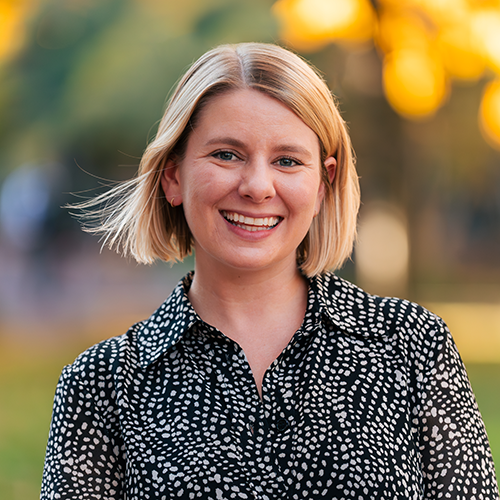 Headshot of a smiling person with short blonde hair, wearing a black and white patterned shirt, outdoors with a blurred background.