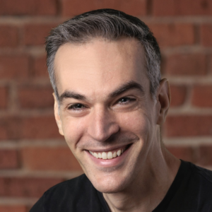 Headshot of a smiling person with short dark hair, wearing a dark shirt in front of a brick wall background.