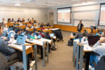 A classroom setting featuring a person giving a presentation to a large group of students. The students are seated with laptops open, engaging with the lecture.