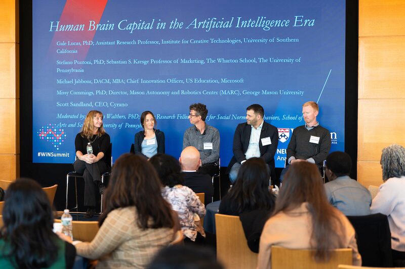 A panel discussion with five speakers seated in front of a backdrop displaying the topic "Human Brain Capital in the Artificial Intelligence Era." An audience is listening attentively.