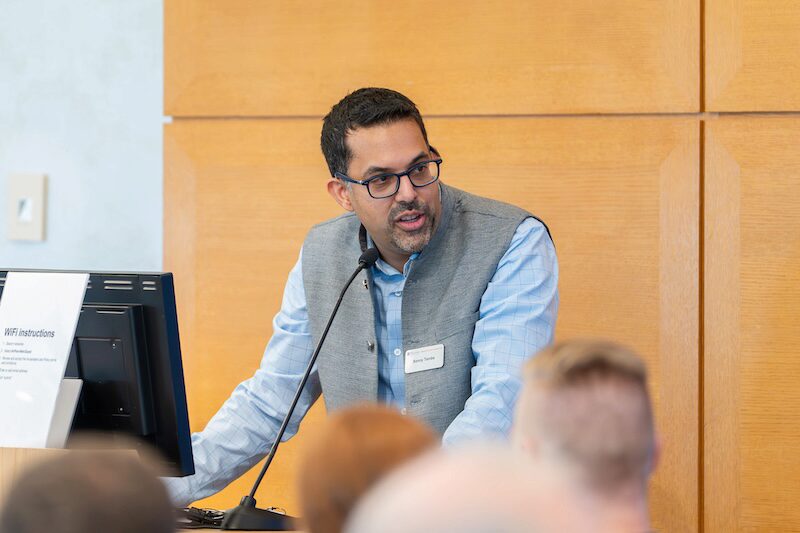 A person speaking at a podium with a microphone, wearing a blue shirt and gray vest, in front of an audience and a computer screen.