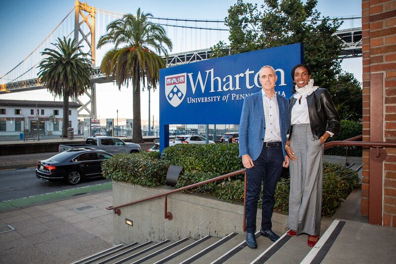 A small group stands in front of a Wharton School of the University of Pennsylvania sign. The background features a bridge and palm trees.