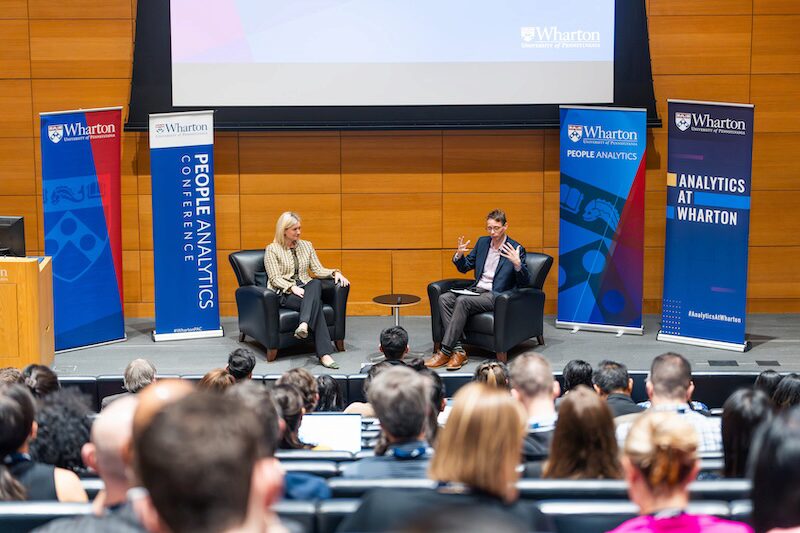 A conversation between two people in armchairs on a stage at Wharton People Analytics Conference, with an audience seated in front.