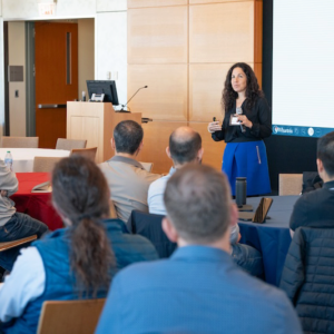 A person is giving a presentation to an audience in a conference room. The presenter is holding a device, and the audience is seated at tables.