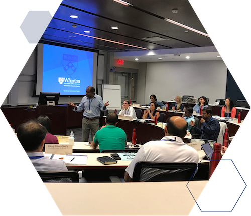 Waiai Corporate Partner Single Image A person is speaking to a small group in a classroom setting with a Wharton School presentation on a screen. Audience members are seated at desks with notebooks and bottles.