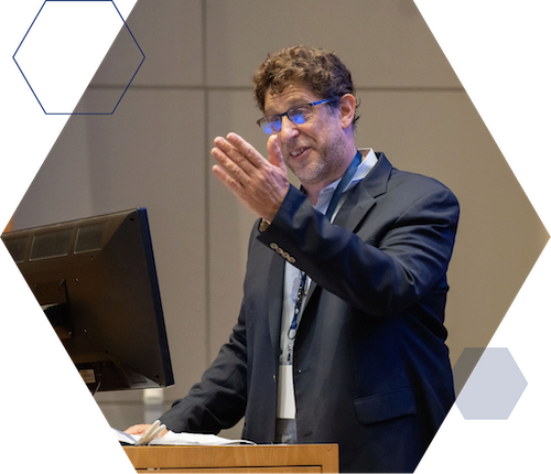 Waiai Faculty Research Partner Single Image A person in a suit gesturing while speaking at a podium with a computer monitor, possibly at a conference or lecture.