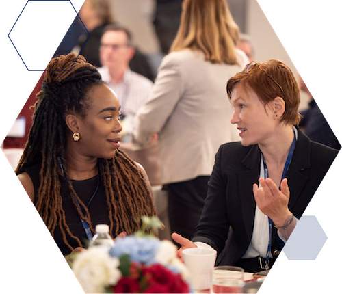 Waiai Individual Partner Single Image A small group of people is engaged in a discussion around a table with drinks and flowers. Two individuals in the foreground are conversing, one with long braided hair and the other with short hair.