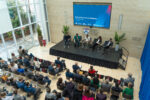 A wide shot from above shows audience members watching a panel of speakers on stage at the Wharton AI Governance Workshop