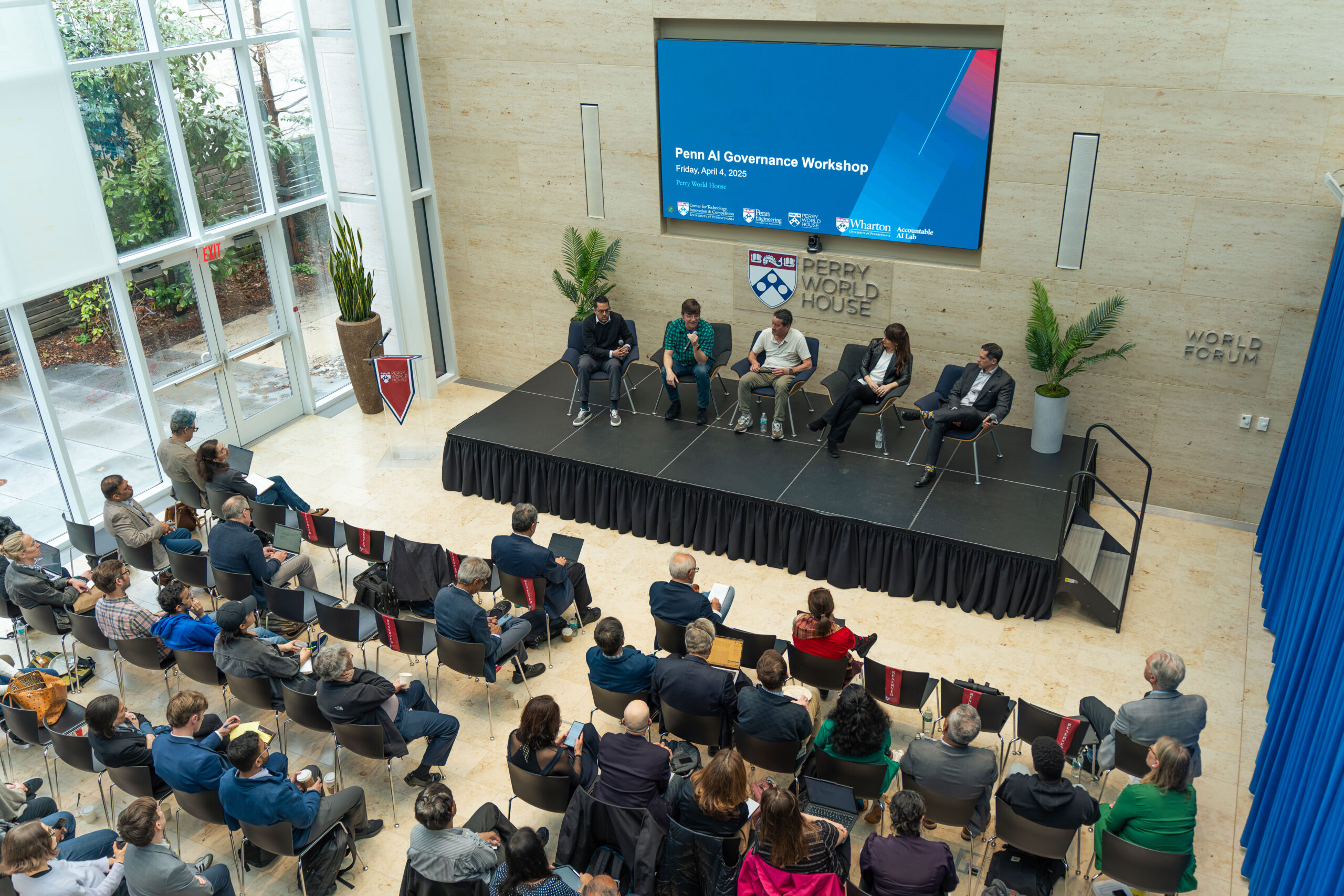 AI Governance 3 A wide shot from above shows audience members watching a panel of speakers on stage at the Wharton AI Governance Workshop