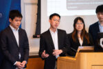 A small group of people in formal attire standing near a podium, with one person speaking. A screen with text is visible in the background.