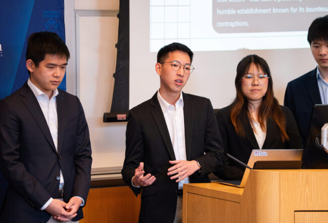 A small group of people in formal attire standing near a podium, with one person speaking. A screen with text is visible in the background.