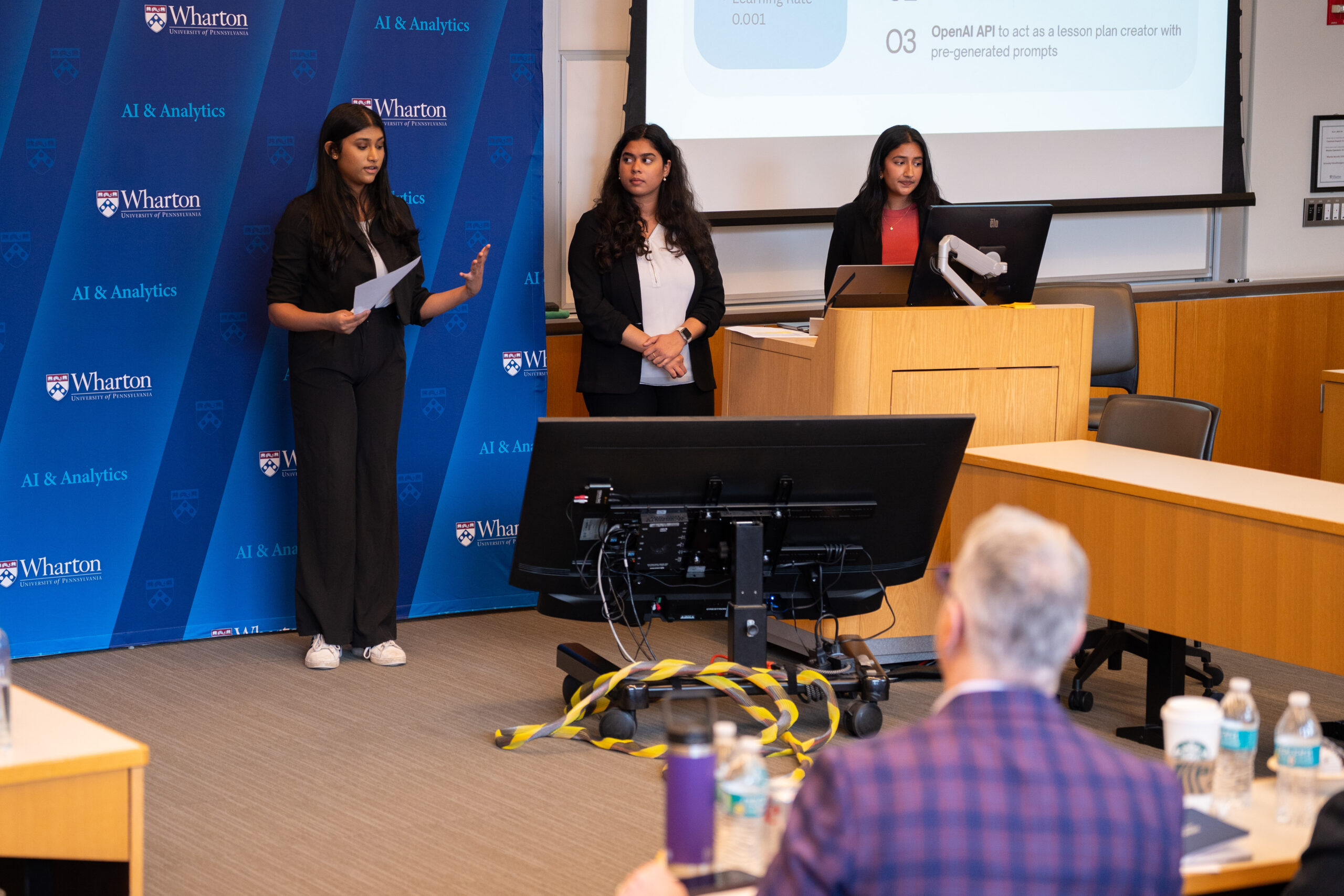 Team M&Ms Presenting A group of individuals is giving a presentation in a classroom setting. A large display screen is visible, and there is a Wharton School banner in the background. The audience includes a person with gray hair wearing a plaid jacket, and bottles of water and