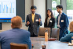 A small group of people in formal attire are giving a presentation. One person is speaking, with a chart displayed on a screen behind them. Others in the room are seated, listening attentively.