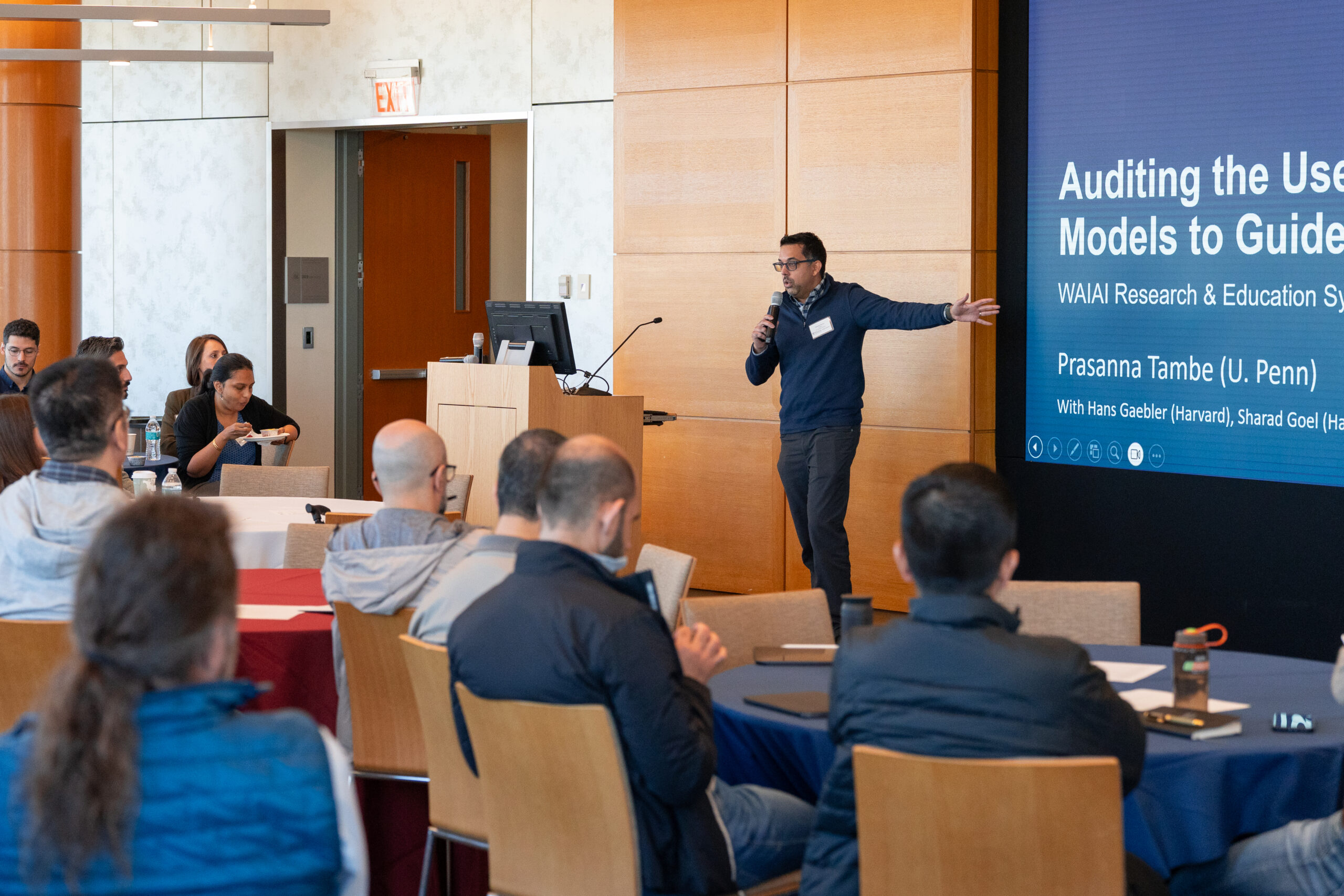 A person is giving a presentation to an audience in a conference room. The screen displays "Auditing the Use of AI Models to Guide..." with names and affiliations. The audience is seated at tables, listening attentively.