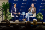 Two people are engaged in a discussion on stage at a Wharton event. They are seated in chairs, with plants and branded banners in the background.