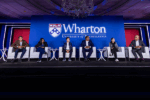 A panel of six people seated on stage at a Wharton event at the University of Pennsylvania, under a large screen displaying the Wharton logo.