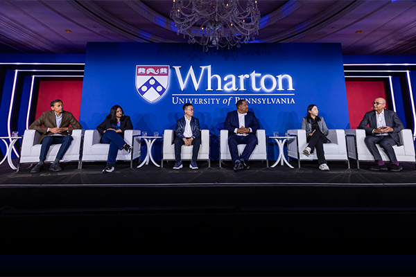 A panel of six people seated on stage at a Wharton event at the University of Pennsylvania, under a large screen displaying the Wharton logo.