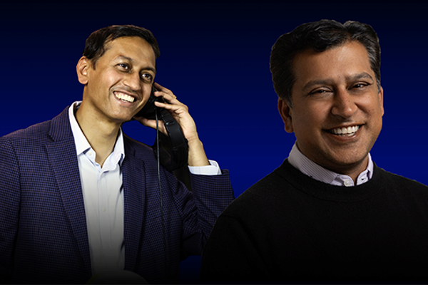 Two people are smiling, one holding a phone. They stand against a dark blue background.