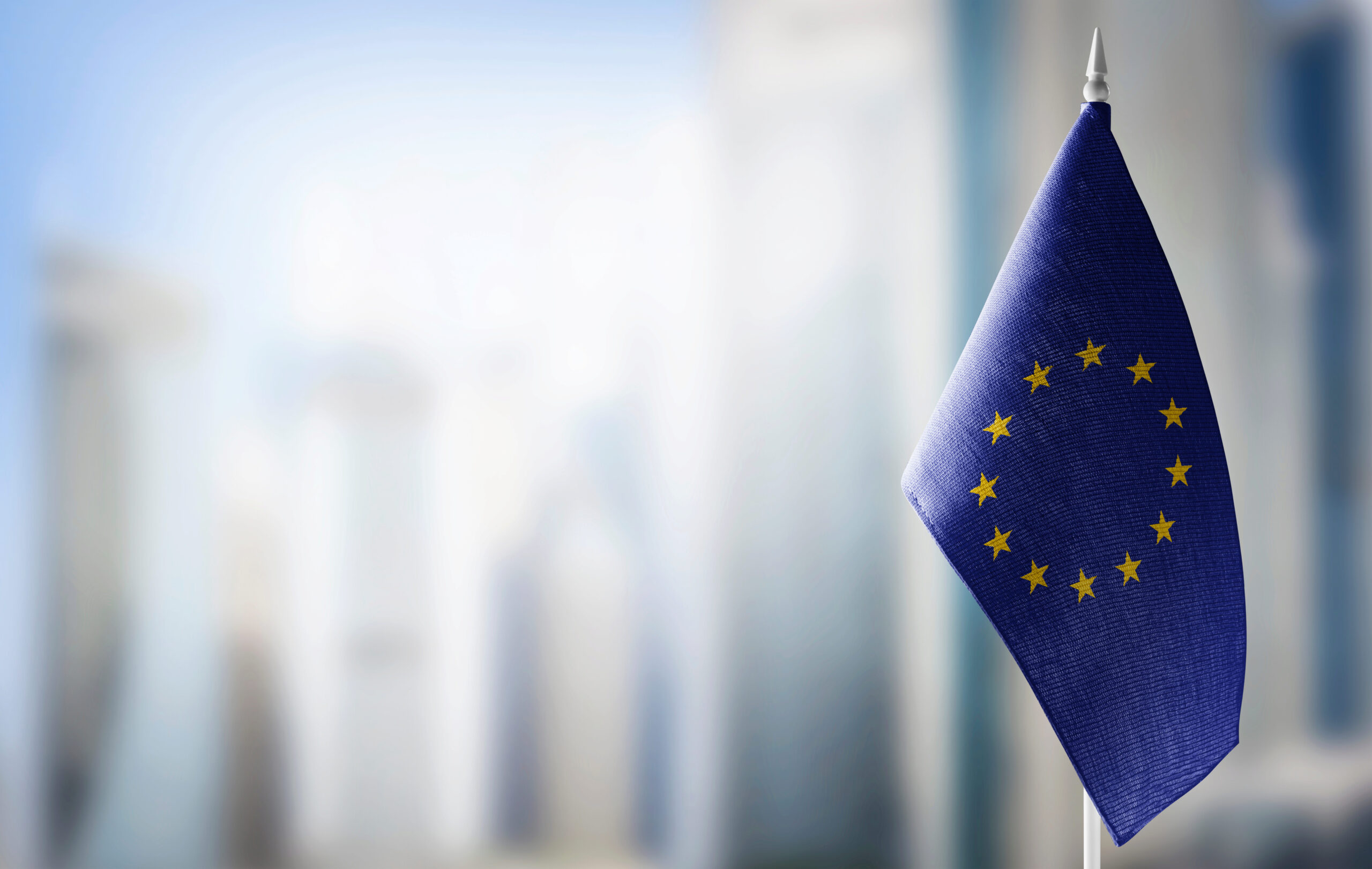 A European Union flag displaying yellow stars in a circle on a blue background, set against a blurred cityscape.