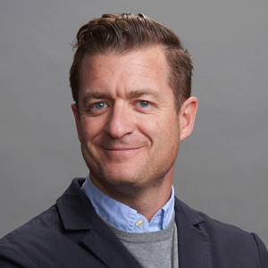 Brett Leary 300x300 Headshot of a smiling person wearing a suit jacket over a sweater and shirt, with a neutral gray background.