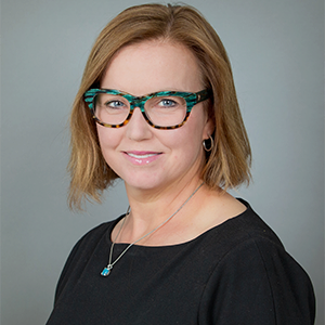 Jen Spada 300x300 Headshot of a person with shoulder-length hair, wearing glasses and a black top, against a neutral background.