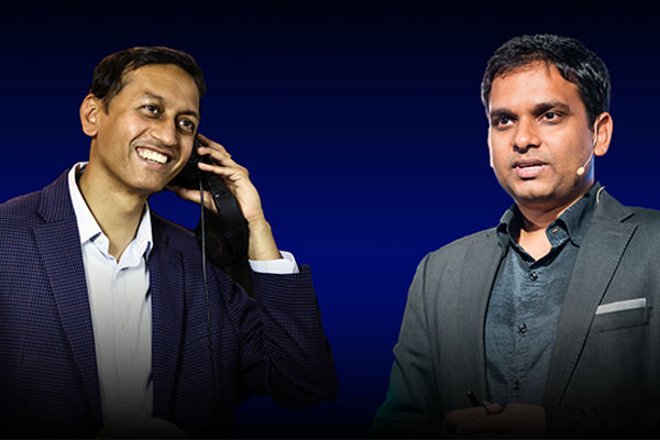 Two people in suits are shown against a blue background, one smiling and holding a phone, the other speaking with a focused expression.