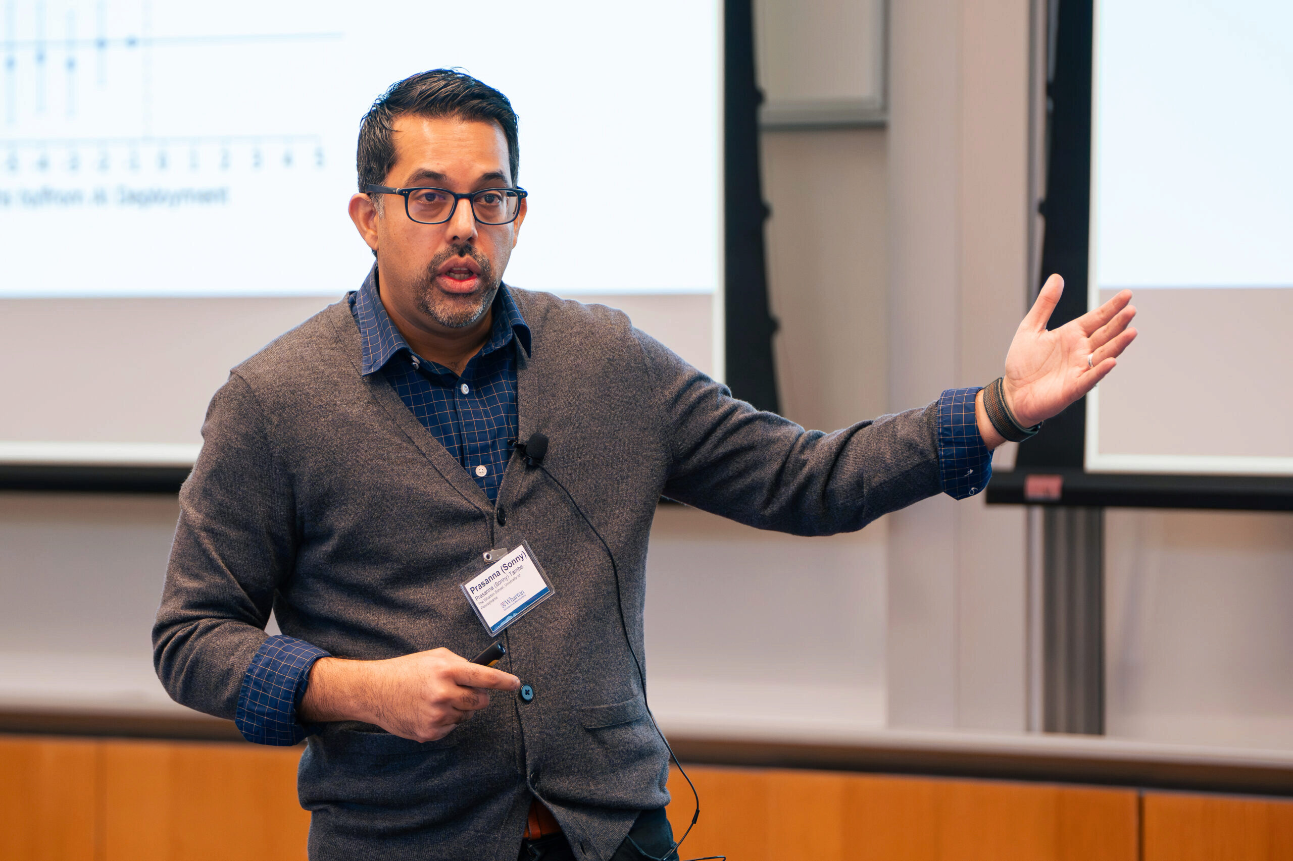 A person giving a presentation, gesturing towards a screen with charts. They wear glasses, a gray cardigan, and a name tag.