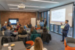 A group of people in a meeting room, some seated at tables and others standing near a projector screen. The screen displays a presentation.