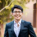 Headshot of a person smiling, wearing glasses and a suit with a white shirt; background features blurred greenery.
