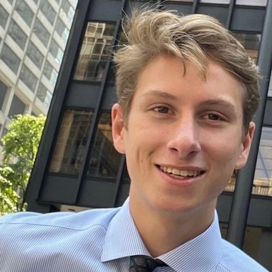 Headshot of a person with short hair, wearing a striped shirt and tie, smiling, with a backdrop of urban buildings.