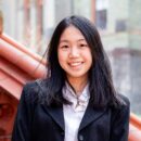 Headshot of a person smiling, wearing a black jacket over a white shirt, with an urban background.