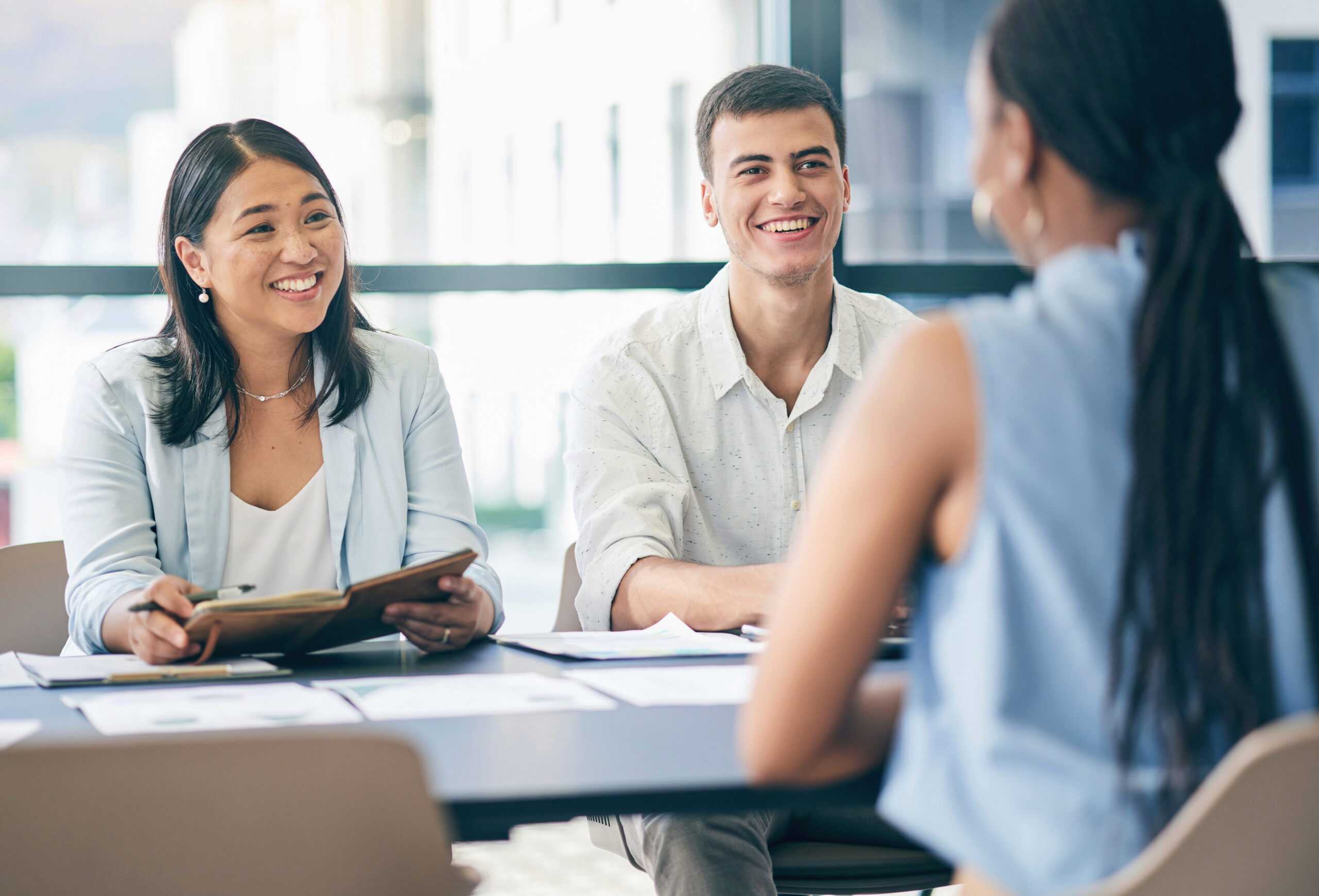 Three people sitting around a table having a meeting, with two facing the camera and one with their back turned. They appear engaged and smiling.