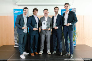 A group of people standing on a stage, holding awards and a device. They are dressed in formal attire. The setting is Tangen Hall at the University of Pennsylvania.